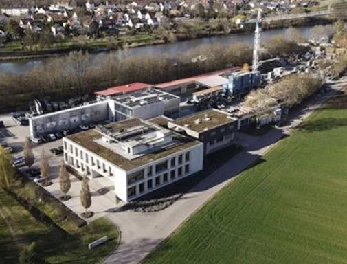Luftbild des Betriebsgeländes: Verwaltungsgebäude und Hallen mit Bauhof, dahinter der Fluss.